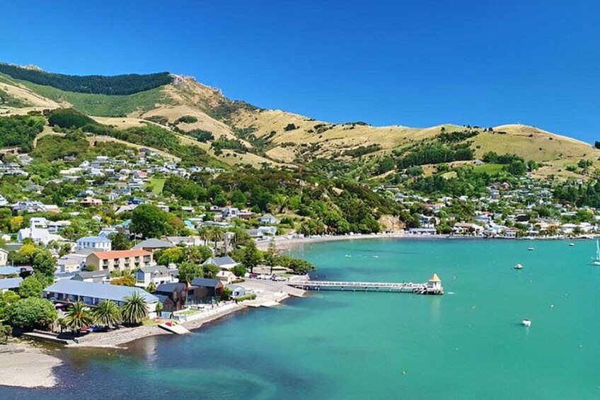 A coastal town with a pier extending into turquoise waters, surrounded by hills and residential buildings. 1