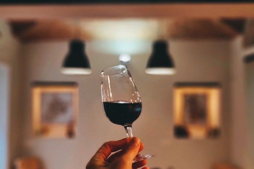 A hand holding a wine glass with a dark liquid, possibly wine, in a room with pendant lights and framed pictures on the wall. 2