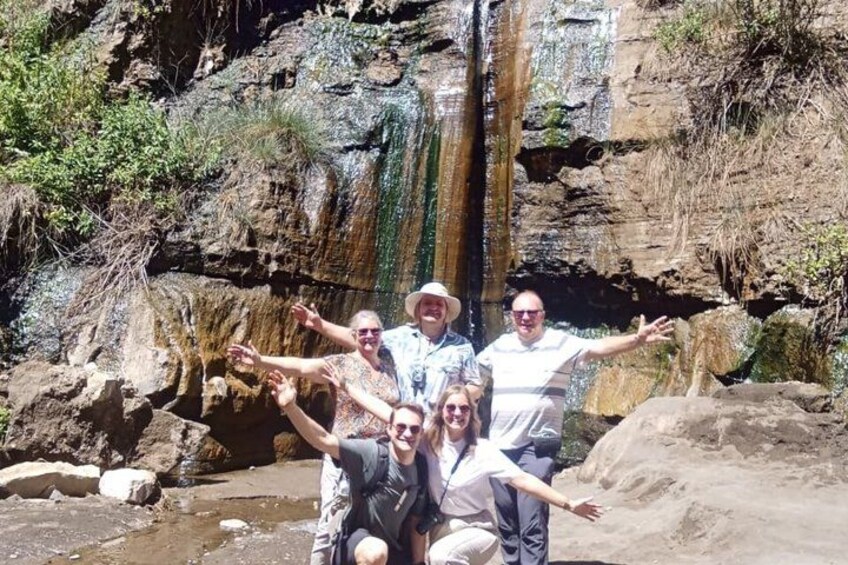 A group of people posing in front of a waterfall with rocky terrain and sparse vegetation. 3