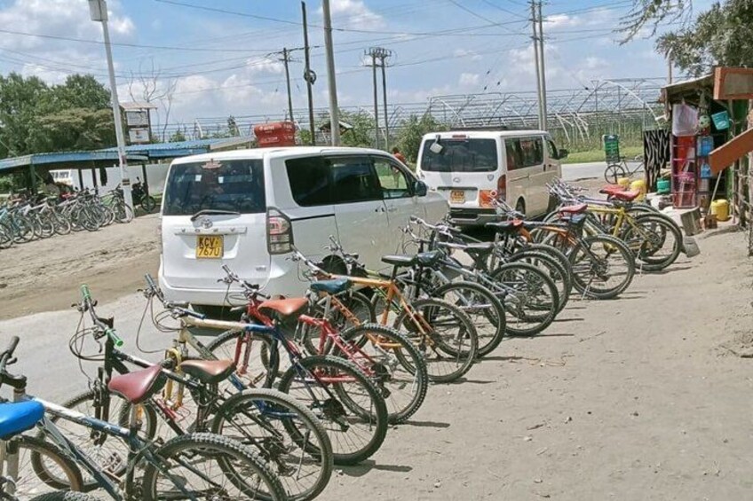 A row of bicycles parked along a dirt road, a white van, and a small building with a blue roof. 4
