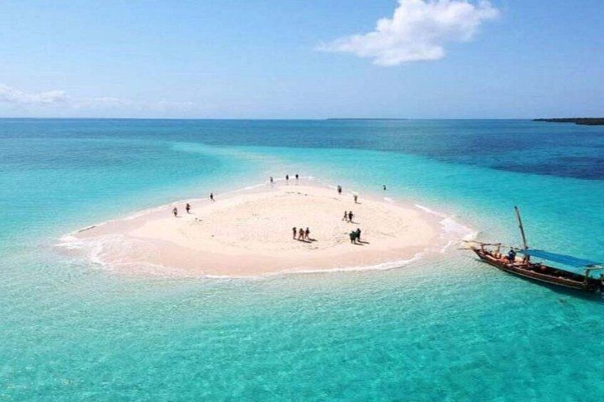 A small island with a sandy beach surrounded by turquoise water, a boat anchored nearby, and a few people scattered across the beach. 1