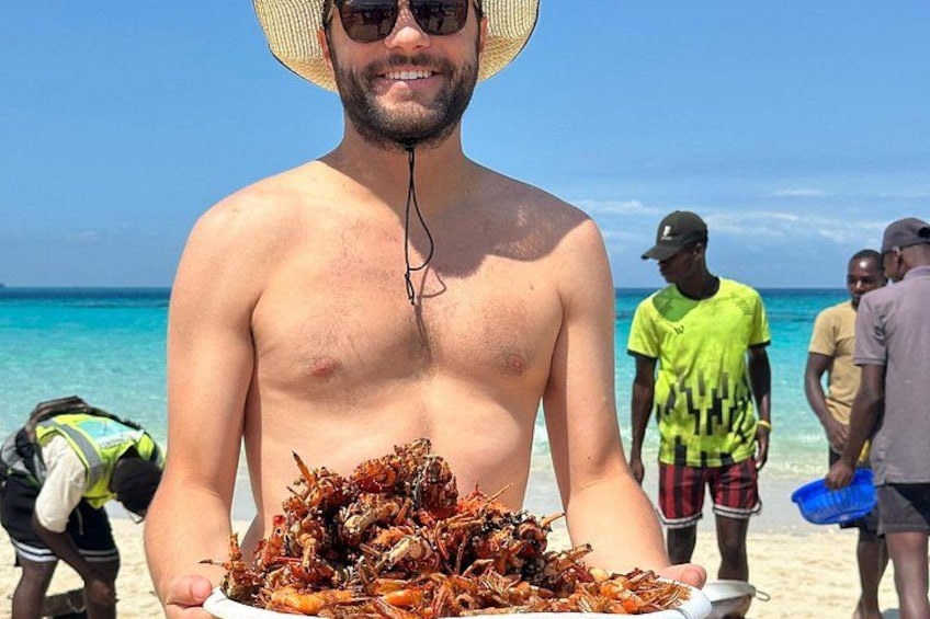 A person holding a plate of cooked seafood at a beach with clear blue water and other people in the background. 5
