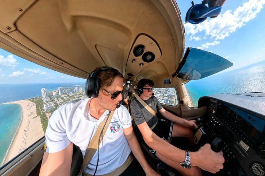 Two people in a cockpit with a view of a coastline and buildings. 1