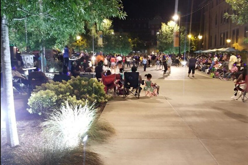 A nighttime outdoor event with people gathered around tables and chairs, illuminated by street lamps. 3
