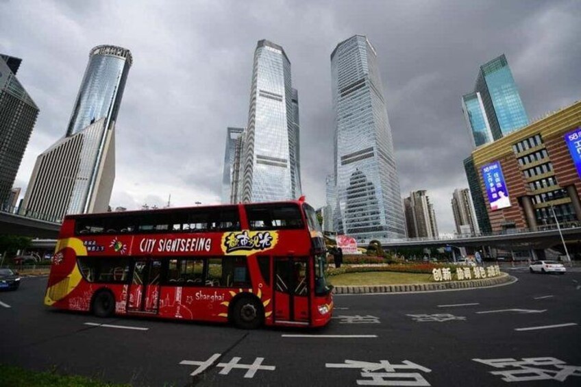 A red double-decker bus with 'CITY SIGHTSEEING' written on it, parked in a city intersection with tall buildings in the background. 1