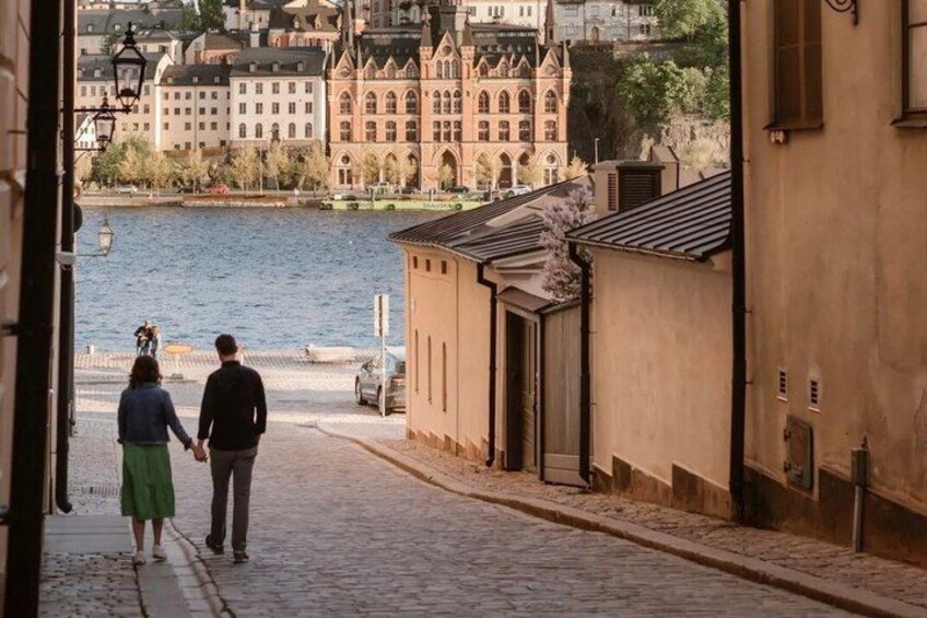 A couple walking down a cobblestone street, a body of water, and historic buildings in the background. 2