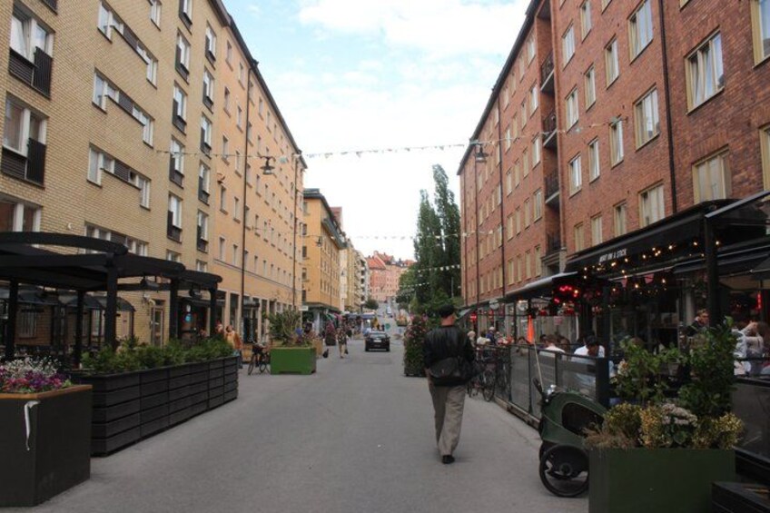 A pedestrian street lined with buildings, outdoor seating areas, and planters with flowers. 5