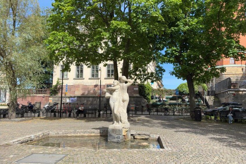 A statue in a square with cobblestone pavement, surrounded by trees and a building in the background. 3