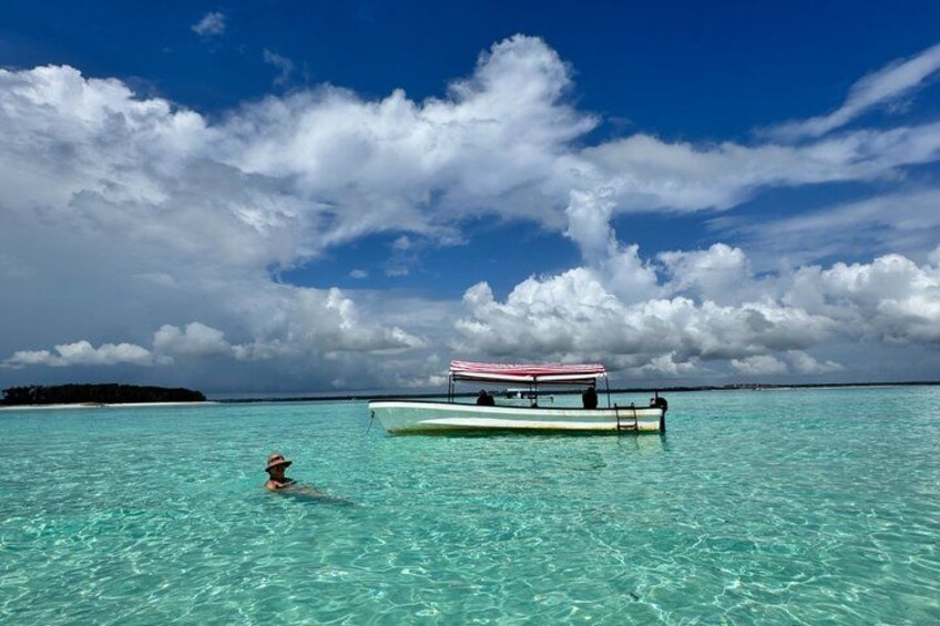 A person swimming in clear turquoise water, a small boat with a striped canopy, and a distant island under a partly cloudy sky. 2
