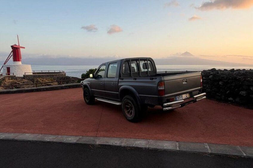 A pickup truck parked on a red surface near a lighthouse and rocky shoreline. 1
