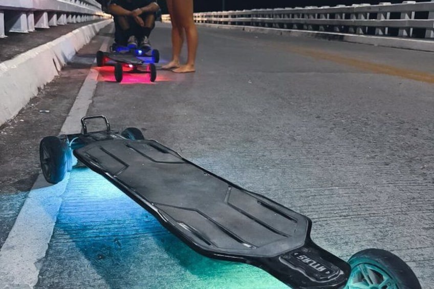 A black electric skateboard on a road with blue and green lights, a person in the background, and a railing. 4