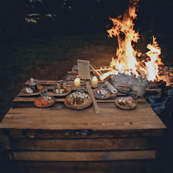 A wooden table with snacks and a sign, set against a campfire. 5