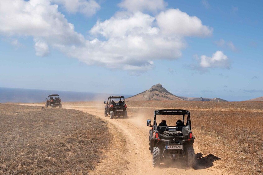 Three off-road vehicles on a dirt road in a dry, open landscape with a mountain in the background. 1