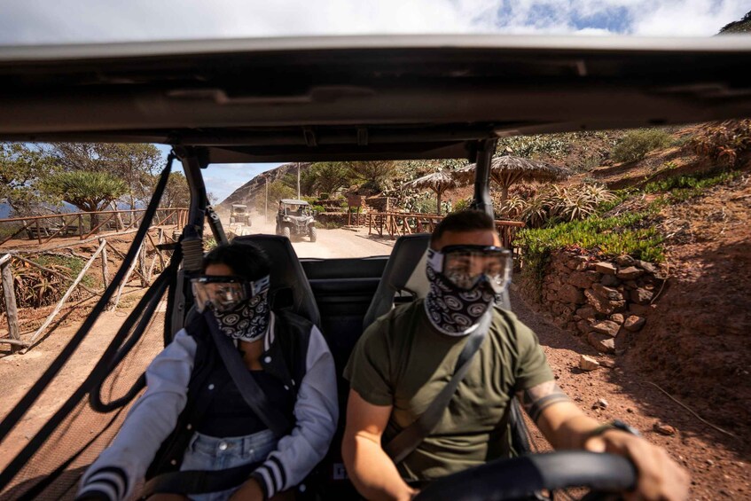 Two people driving a vehicle on a dirt road with a rocky hillside and sparse vegetation in the background. 3