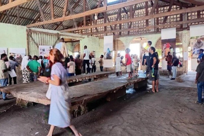 A crowded indoor space with a high ceiling and wooden beams, featuring a central stage and people gathered around. 3