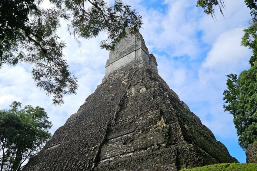 A large, ancient stone pyramid with a narrow top, surrounded by trees and a clear sky. 1