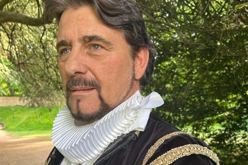 A person in historical attire with a ruffled collar and ornate details, standing in a lush green park with trees and a pathway. 1