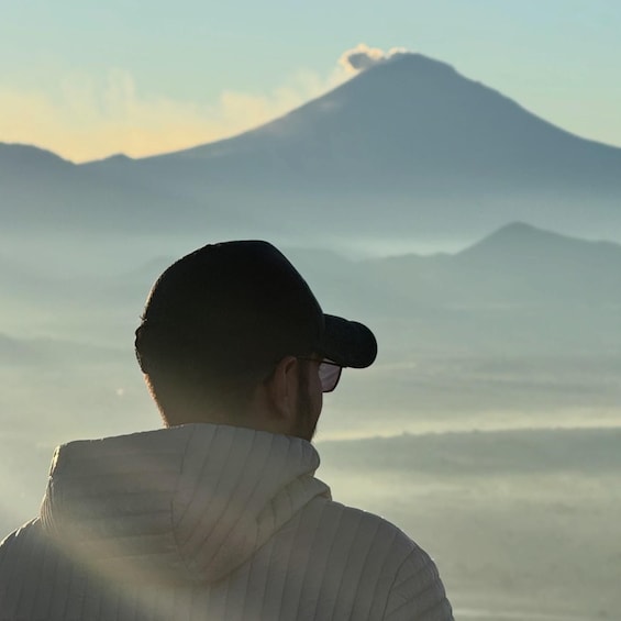 A person wearing a white jacket and a cap, looking at a mountain with a cloud on top. 3