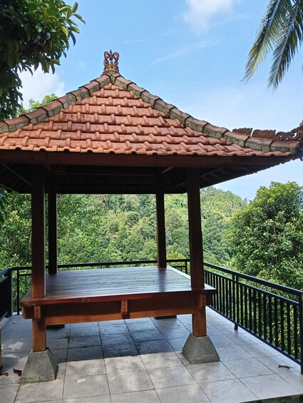A gazebo with a tiled roof and a wooden floor, surrounded by a metal railing, overlooking a lush green forest. 2