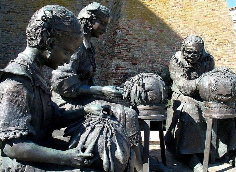 A bronze statue of three figures, one seated and two standing, in an outdoor setting with a brick wall in the background. 3