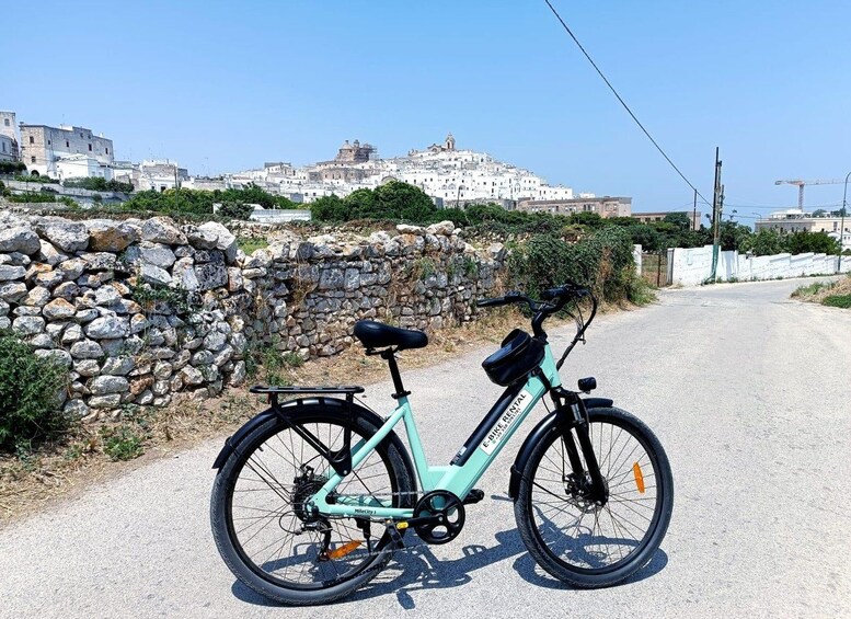 A teal E-Bike parked on a road with a stone wall and buildings in the background. 4