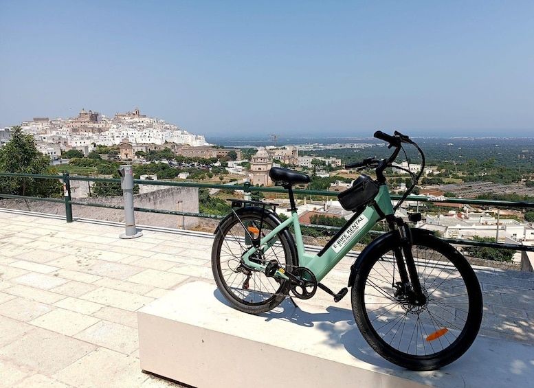 A green electric bike parked on a tiled platform overlooking a coastal cityscape with historic buildings. 1