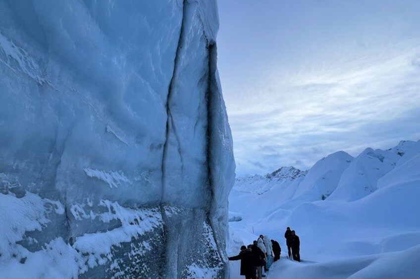 A group of people walking on snow-covered terrain with a large ice formation in the background. 4