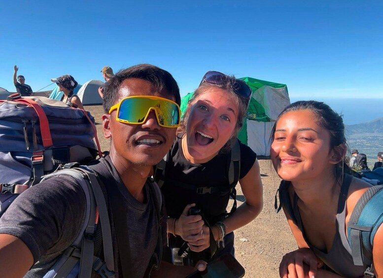 Three people with backpacks, a tent, and a mountainous landscape in the background. 1
