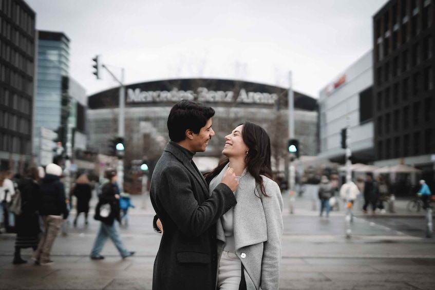 A couple embracing in a city square with a Mercedes-Benz Arena in the background. 3