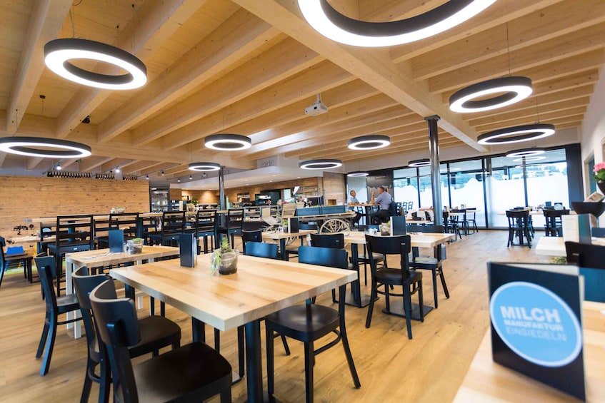 A modern cafeteria with wooden tables and chairs, circular ceiling lights, and a visible sign for 'MILCH'. 2