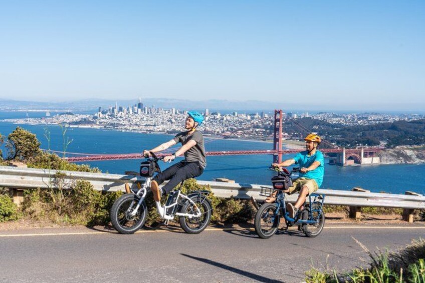 Two people riding bicycles on a road with a city skyline and a bridge in the background. 3