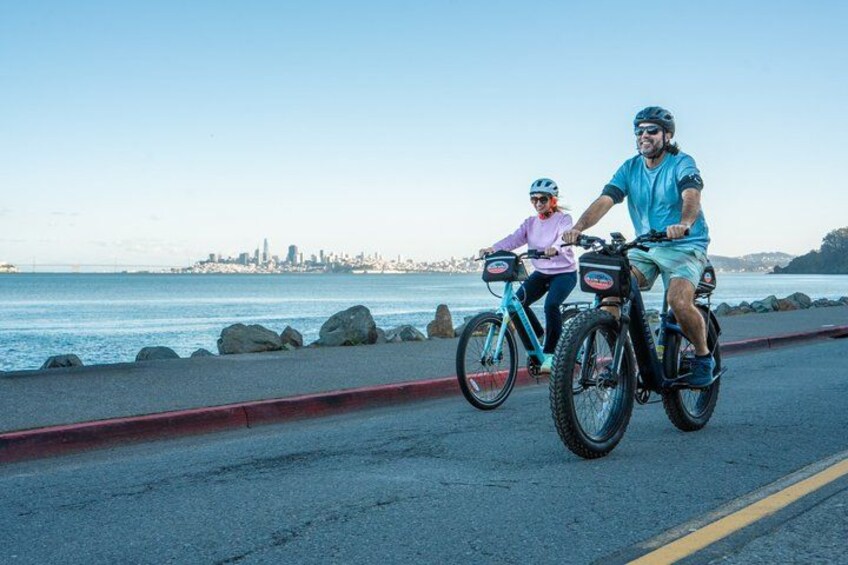 Two people riding bicycles on a coastal road with a city skyline in the background. 2