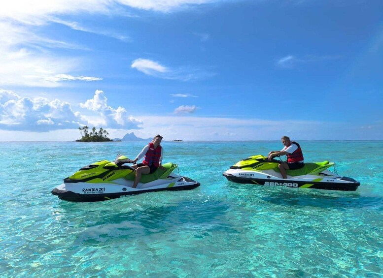 Two people jet skiing on clear turquoise water with a tropical island and mountains in the background. 1