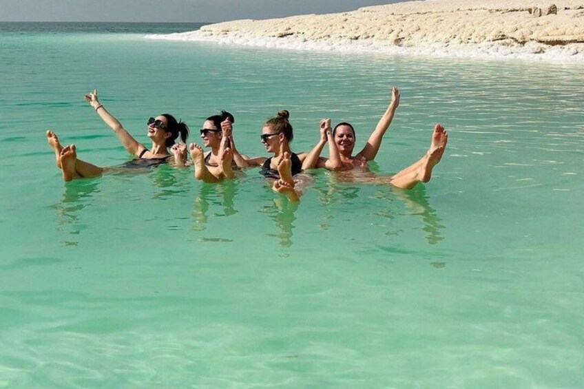 Four people floating in clear turquoise water with a sandy beach and rocky outcrops in the background. 1