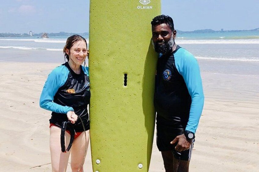 Two people standing on a sandy beach with a green surfboard between them. 2