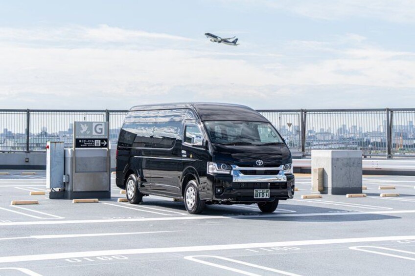 A black van parked in a lot with a cityscape and an airplane in the background. 3