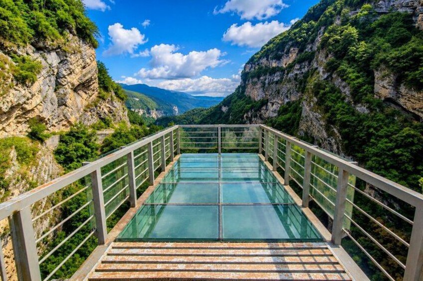 A glass bridge with metal railings, surrounded by lush greenery and rocky cliffs. 5