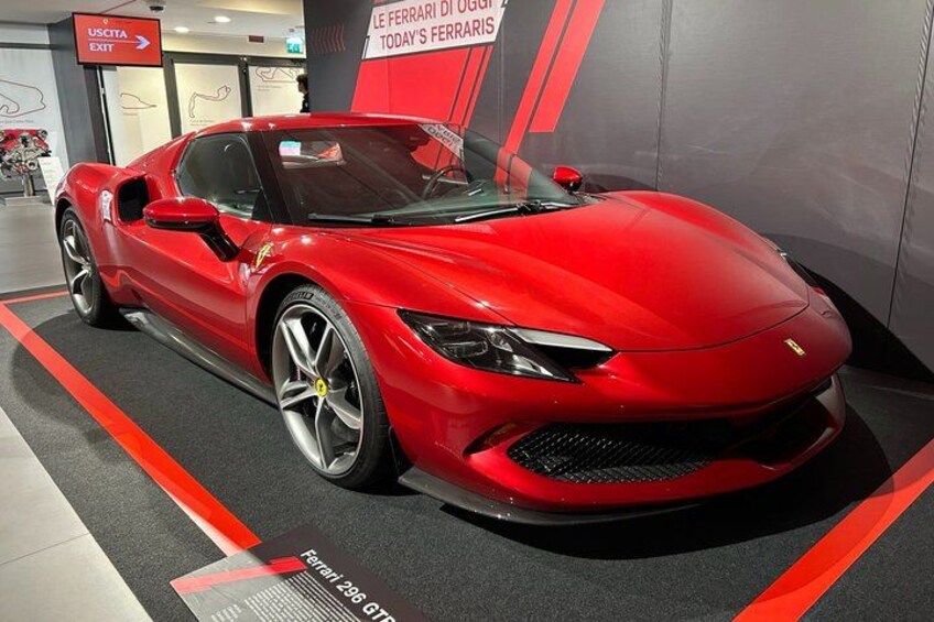 A red sports car on display with a sign reading 'LE FERRARI 296 GT3' and an exit sign in the background. 2