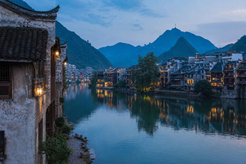 A riverside town with traditional architecture, a calm river, and mountains in the background. 1