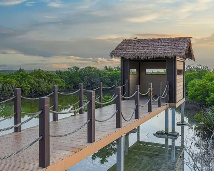 A wooden walkway with a thatched-roof structure, surrounded by water and greenery. 2