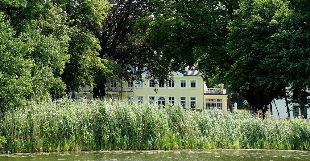 Talo, jonka ympärillä on korkeita puita ja jonka lähellä on vesialue, jossa kasvaa ruokoa. 1