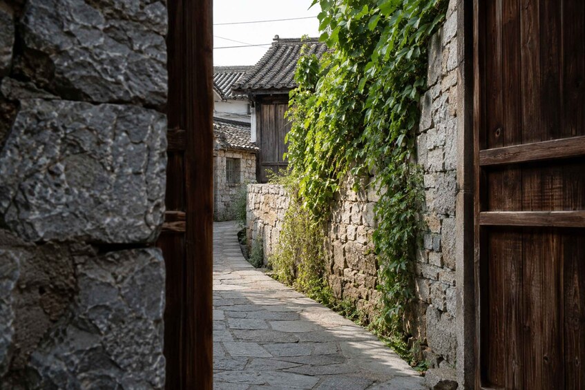 A narrow stone pathway flanked by stone walls and wooden doors, leading to traditional buildings with tiled roofs. 2