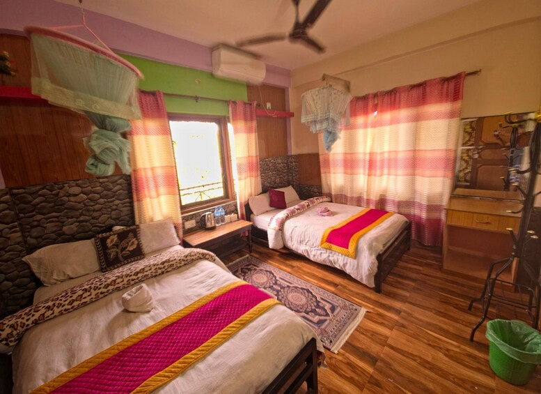 A room with two beds, a ceiling fan, a window with curtains, a green bucket, and a wooden desk. 3