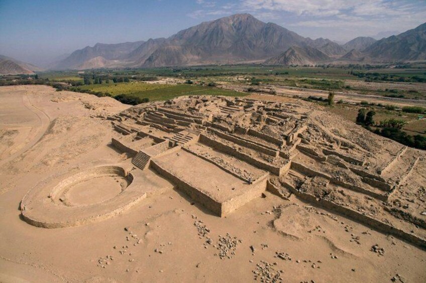 An ancient archaeological site with terraced structures and a large circular area, set against a mountainous backdrop. 1