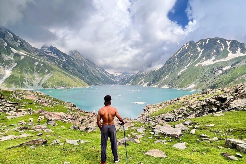 A man standing on a grassy hill overlooking a lake with mountains in the background. 2