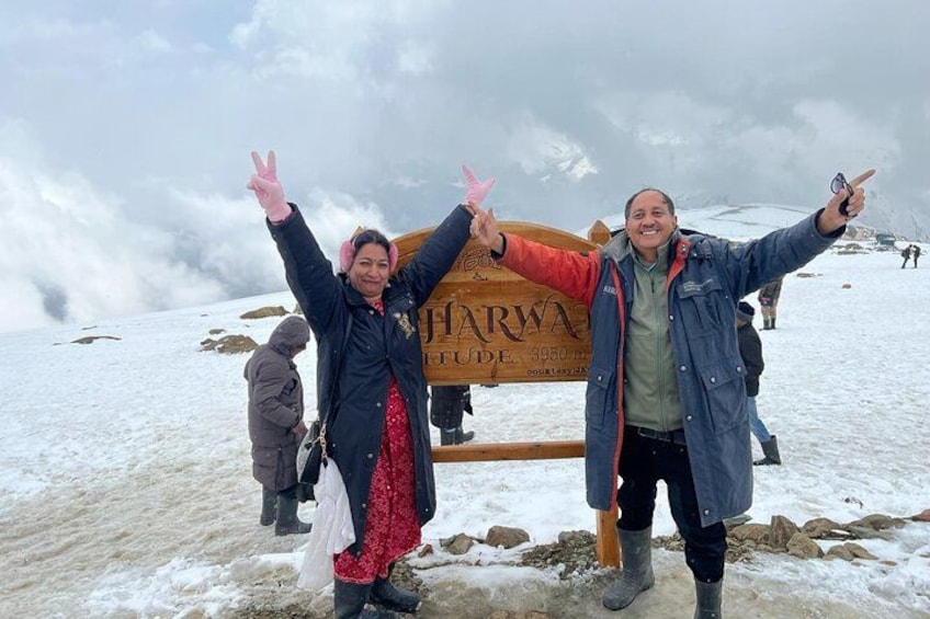 Two people standing on a snowy mountain with a sign that reads 'JHARWA'. 1