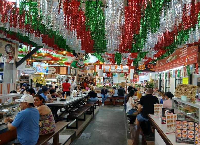 A bustling indoor market with people dining at tables, decorated with red, green, and white hanging ornaments. 3
