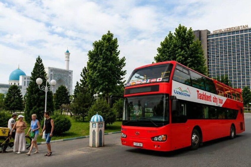 A red double-decker bus with 'Tashkent city tour' written on it, parked on a street with people walking around, trees, and a building with a dome in the background. 1