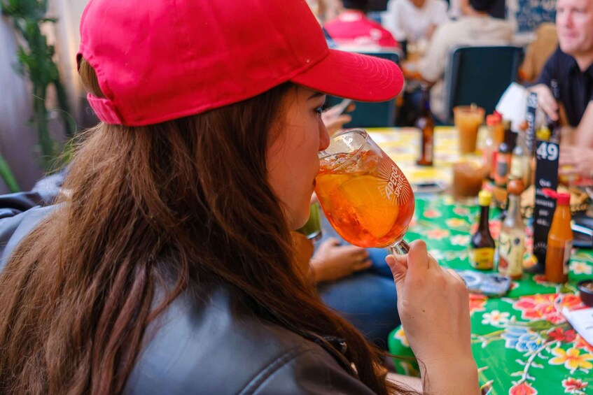 Una persona con gorra roja, sosteniendo un vaso con una bebida, sentada en una mesa con varias botellas y un mantel con motivos florales. 3