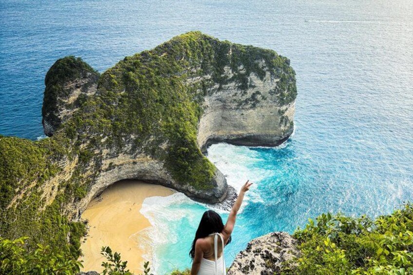 A person standing on a cliff overlooking a beach with a large rock formation and turquoise waters. 3